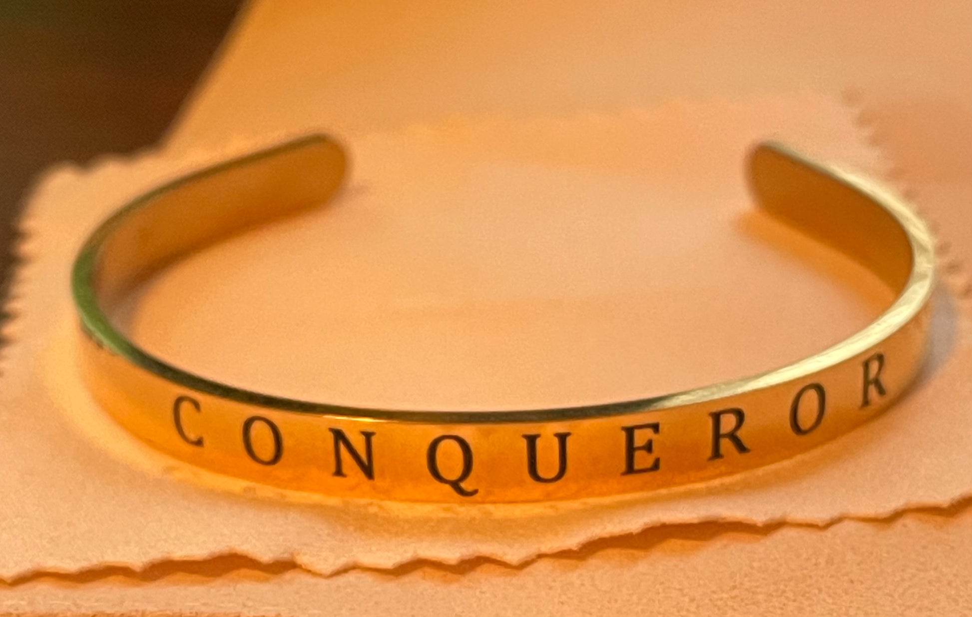 Gold bracelet with 'CONQUEROR' engraving on a beige background