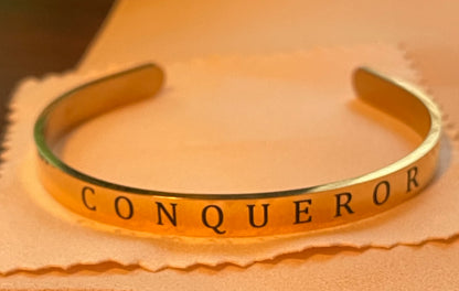 Gold bracelet with 'CONQUEROR' engraving on a beige background